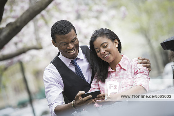 Stadtleben im Frühling. Jugendliche im Freien in einem Stadtpark. Ein Paar nebeneinander  den Arm um ihre Schultern gelegt  ein Smartphone betrachtend und lächelnd.