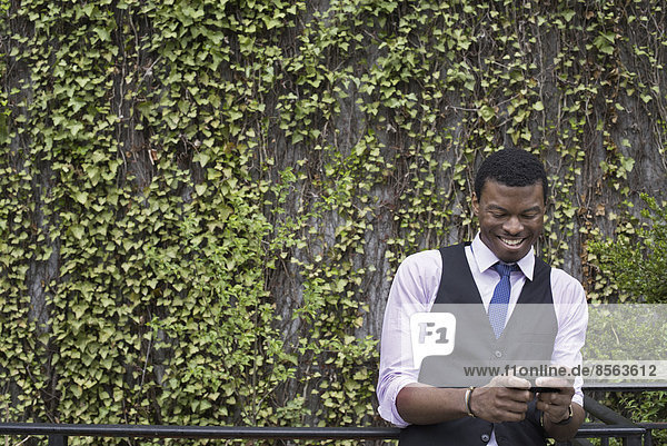 City life in spring. City park with a wall covered in climbing plants and ivy. A young man in a waistcoat  shirt and tie checking his phone.