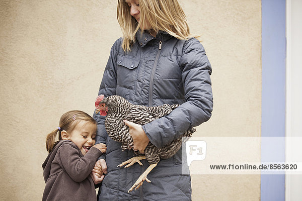 Eine Frau in grauem Mantel  die ein schwarz-weißes Huhn mit rotem Hahnenkamm unter einem Arm hält. Ein junges Mädchen neben ihr  das die andere Hand hält.