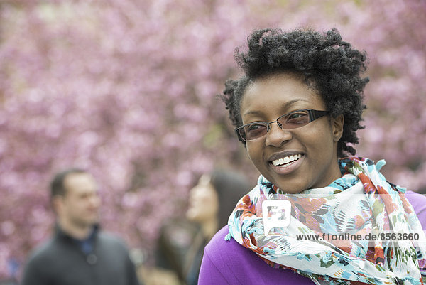 A group of people under the cherry blossom trees in the park. A young woman smiling  wearing a purple shirt and floral scarf.