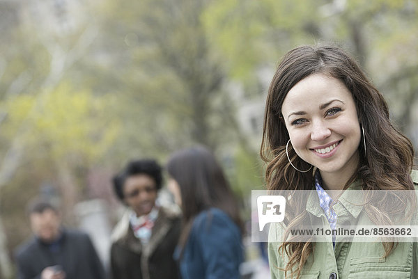 Eine Gruppe von Menschen im Park. Eine junge Frau in einem Hemd mit offenem Hals  die lächelt und in die Kamera schaut.