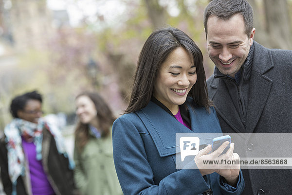 A group of people in a city park. A man in a grey coat  and a woman in a turquoise coat  both looking at a smart phone.