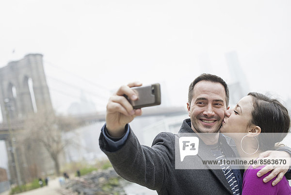 New York Stadt. Die Brooklyn Bridge über den East River. Ein Paar beim Fotografieren mit einem Telefon  ein Selfy von sich selbst.