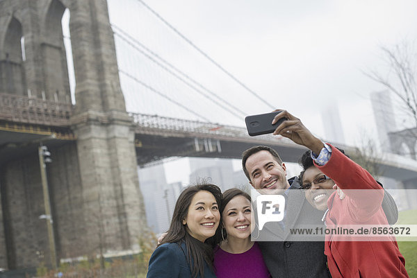 New York Stadt. Die Brooklyn Bridge über den East River. Vier Freunde  die mit einem Telefon ein Foto von sich selbst machen.