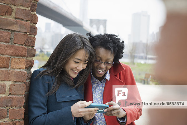 New York City  die Brooklyn Bridge  die über den East River führt. Zwei Frauen  Seite an Seite  schauen auf den Bildschirm eines Smartphones und lachen.