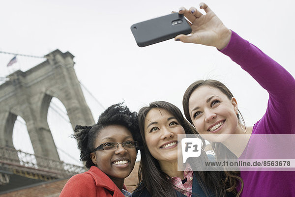 New York City  die Brooklyn Bridge  die über den East River führt. Drei Frauen in einer Reihe  lächelnd  als man ein Foto mit einem Smartphone macht.