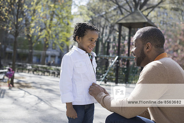 A New York city park in the spring. Sunshine and cherry blossom. A father kneeling and buttoning his son's jacket.
