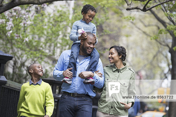 A New York city park in the spring. A family  parents and two boys. A child riding on his father's shoulders.