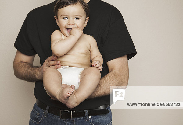 A young boy wearing cloth diapers being held by his father in his arms.