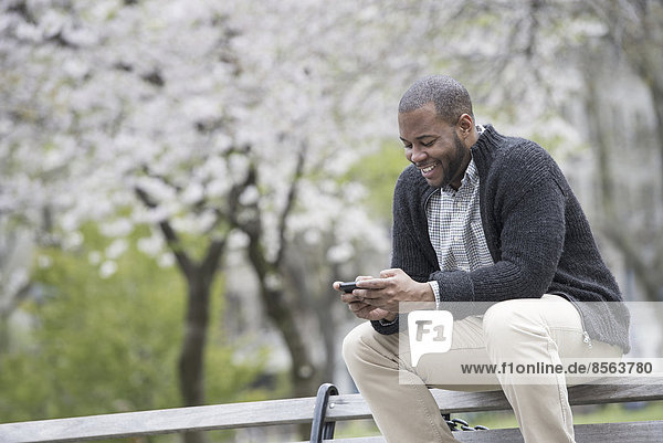 Draußen in der Stadt im Frühling. Ein urbaner Lebensstil. Ein junger Mann  der sein Telefon überprüft und SMS schreibt.
