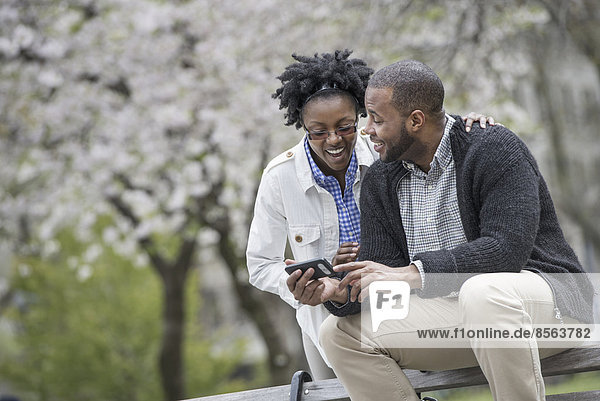 Draußen in der Stadt im Frühling. Ein urbaner Lebensstil. Ein Paar sitzt auf einer Bank  einer hält ein Telefon in der Hand.