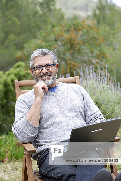 Man sitting in his garden with laptop  smiling and wearing glasses