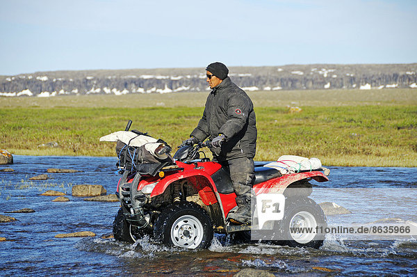 Man of the Inuit people riding a quad bike ATV through a river in the tundra on Victoria Island formerly Holman Island village of Ulukhaktok Northwest Territories Canada