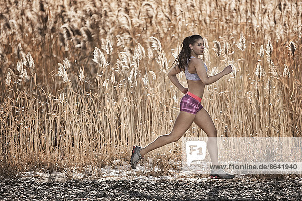Eine Frau rennt durch einen Bestand hoher Gräser  die größer werden  als sie ist. Braun gefärbte Ufergräser mit gefiederten Samenköpfen.