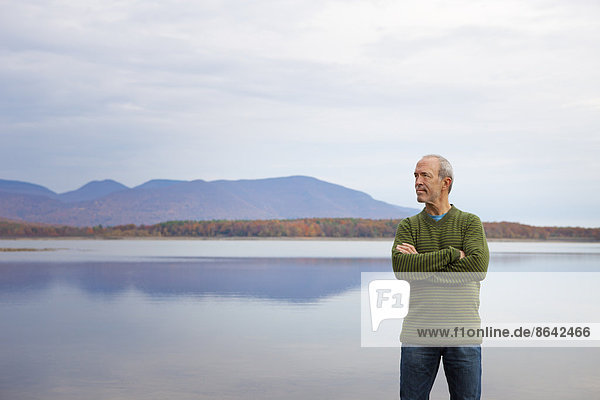 A man with arms folded by the water of a flat calm lake in the mountains.