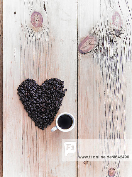 Draufsicht auf einen Haufen gerösteter Bio-Kaffeebohnen in Herzform  die auf einer Tischplatte angeordnet sind. Eine Tasse schwarzer Kaffee.