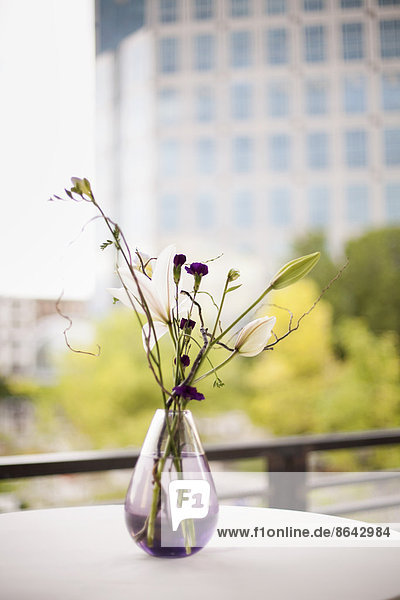 Ein Tisch auf einer Terrasse in der Stadt. Eine Vase mit Blumen. Kleine violette Blumen  sowie weiße Lilien- und Orchideenblüten.