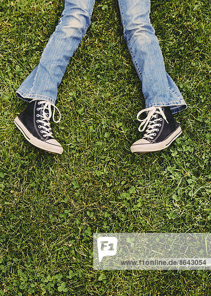 A ten year old girl lying on the grass. Cropped view of her lower legs. Wearing sneakers and faded blue jeans.