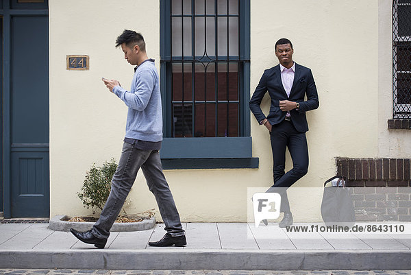Young people outdoors on the city streets in springtime. A man leaning against a wall and one walking past checking his phone.