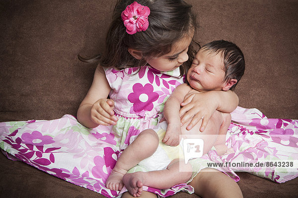 A young girl with long brown hair  with a ribbon hair accessory  in a pink flowered dress holding a newborn baby sibling.