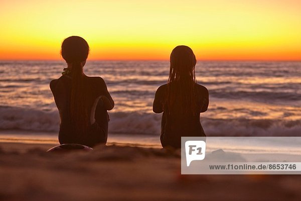 Zwei Mädchen sitzen bei Sonnenuntergang am Strand und schauen aufs Meer.