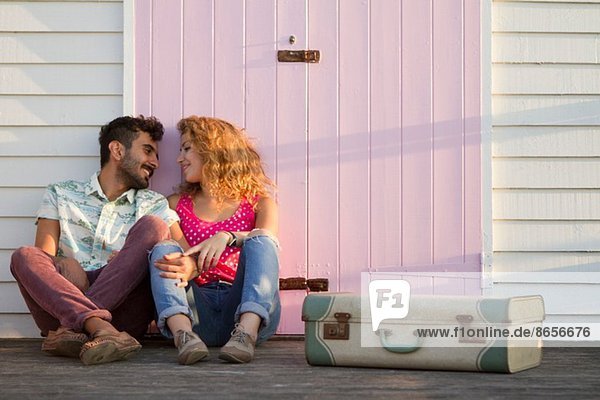 Young couple sitting by beach hut with suitcase