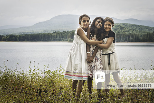 Drei junge Mädchen stehen am Ufer eines Sees und umarmen sich gegenseitig.