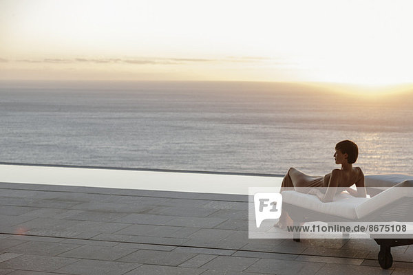 Frau im Kleid auf einem Sessel auf der Terrasse mit Blick auf das Meer bei Sonnenuntergang.