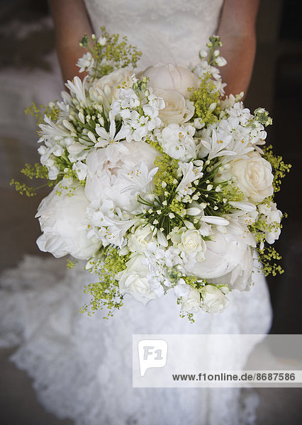 Eine Frau in einem weißen Kleid,  eine Braut mit einem Brautstrauß aus weißen Blumen,  großen weißen Rosen und Pfingstrosen,  mit zarten gelben Blüten und grünen Blättern.