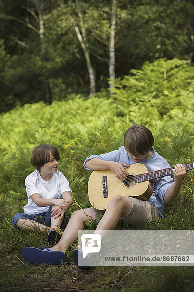 Two boys camping in New Forest. Sitting on the grass  one playing a guitar.