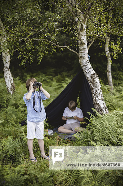 Two boys camping in New Forest. One sitting under a black canvas shelter. One boy looking through binoculars.