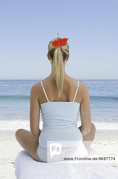 Der Strand. Eine junge Frau sitzt in entspannter Pose auf einem Handtuch im Sand und schaut aufs Meer hinaus.