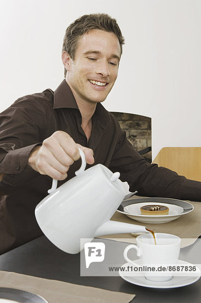 Ein Mann sitzt  gießt sich eine Tasse Kaffee ein und frühstückt.
