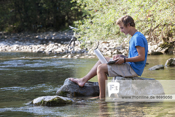 Austria  Salzkammergut  Mondsee  young man with laptop learning at a brook