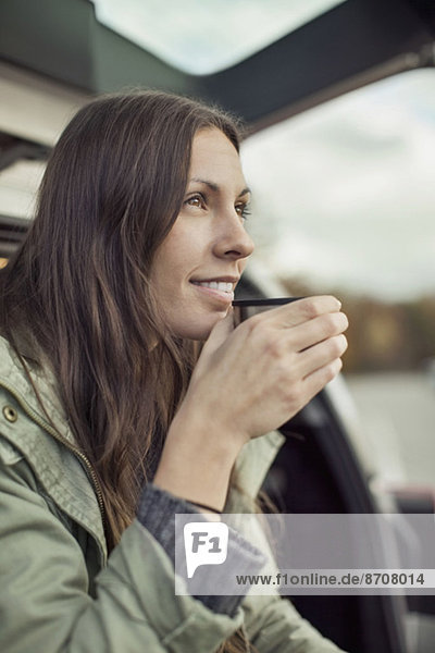 Junge Frau schaut weg und trinkt Kaffee am Kofferraum des Autos.