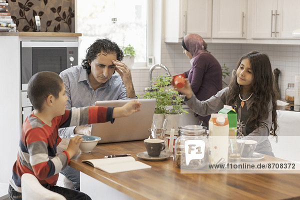 Muslimischer Mann arbeitet am Laptop in der Küche mit Familie beim Frühstücken