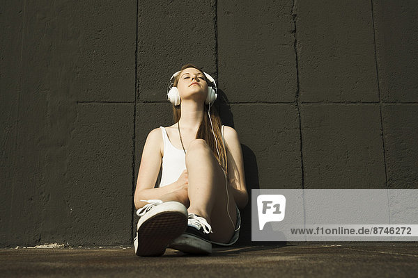 Young woman sitting on ground,  leaning against cement wall,  listening to MP3 player