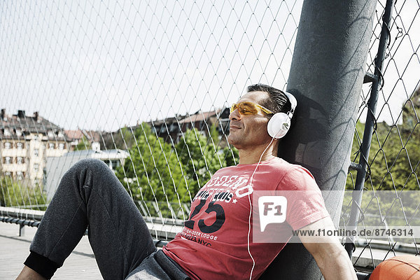 Mature man sitting on outdoor basketball court wearing headphones and listening to music  Germany