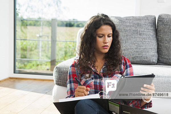 Teenage girl sitting on floor next to sofa  writing in binder and using tablet computer  Germany
