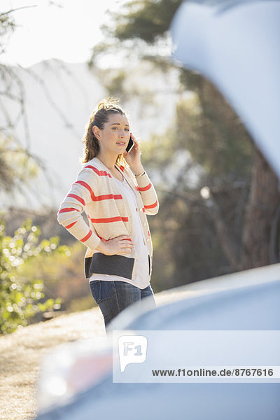 Woman talking on cell phone with automobile hood raised at roadside