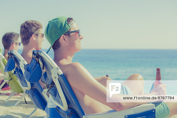 Junge Männer trinken Bier in Liegestühlen am sonnigen Strand