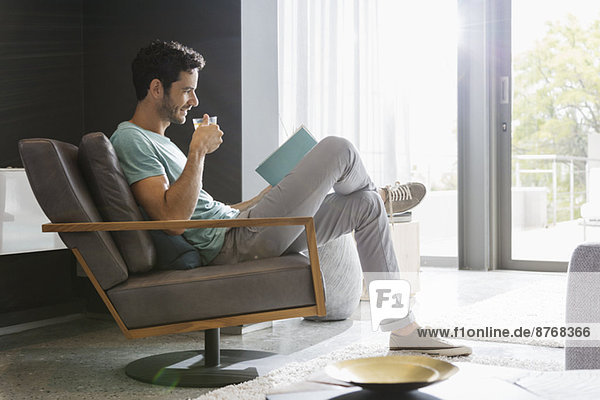Mann trinkt Tee und liest Buch im Wohnzimmer