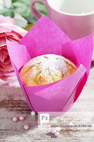 Muffin in rosa Hülle,  Rosenblüten,  Tasse und Zuckerperlen auf Holztisch