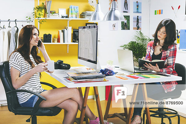 Two female fashion bloggers sitting at desk in the office