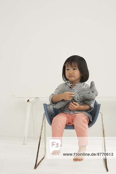 Kleines asiatisches Mädchen mit Plüschtier auf einem Stuhl sitzend
