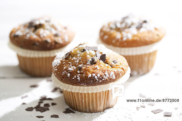 Three muffins in paper cups sprinkled with powdered sugar and chocolate shavings on white wooden table