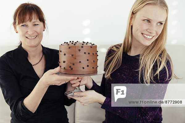 Zwei lächelnde Frauen tauschen Kuchenständer mit Schokoladenkuchen aus.