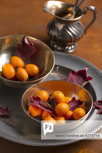 Zwei Metallschalen mit Kumquats,  Schokoladenstücken und roten Herbstblättern auf Metallplatte und Holztisch