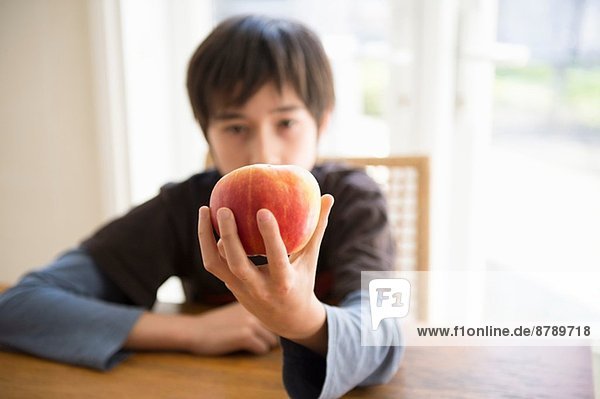 Junge sitzt am Tisch und hält den Apfel vor sich.