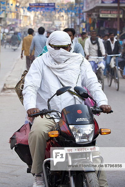 Man driving Hero Honda motorcycle with covered head and face in street scene in city of Varanasi  Benares  Northern India
